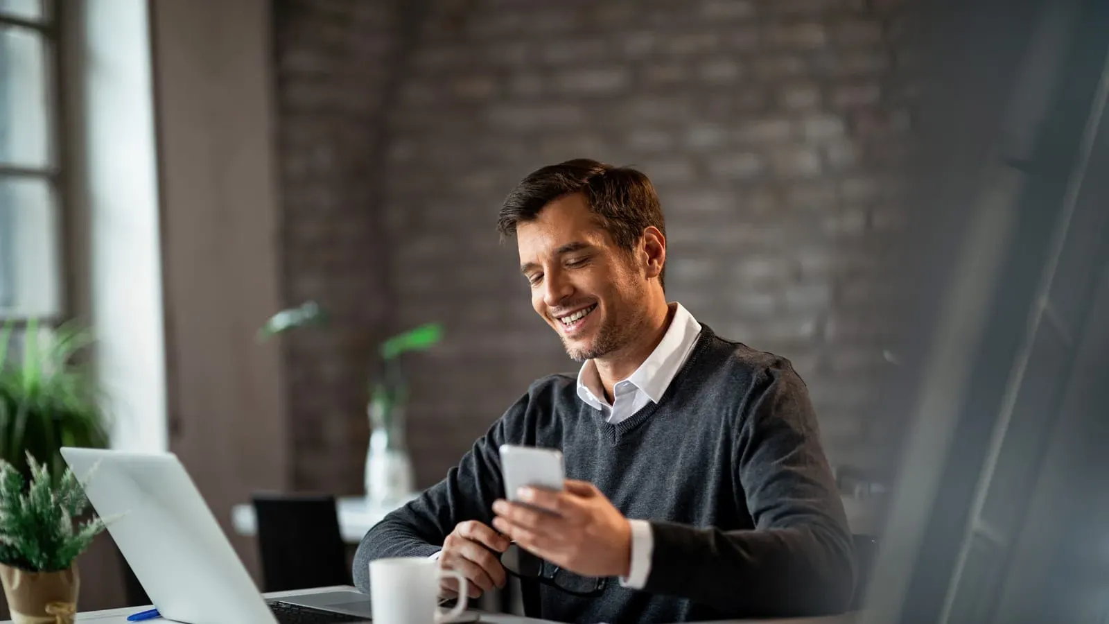 Vendedor sorrindo enquanto responde um cliente no celular, sentado à mesa com o notebook aberto